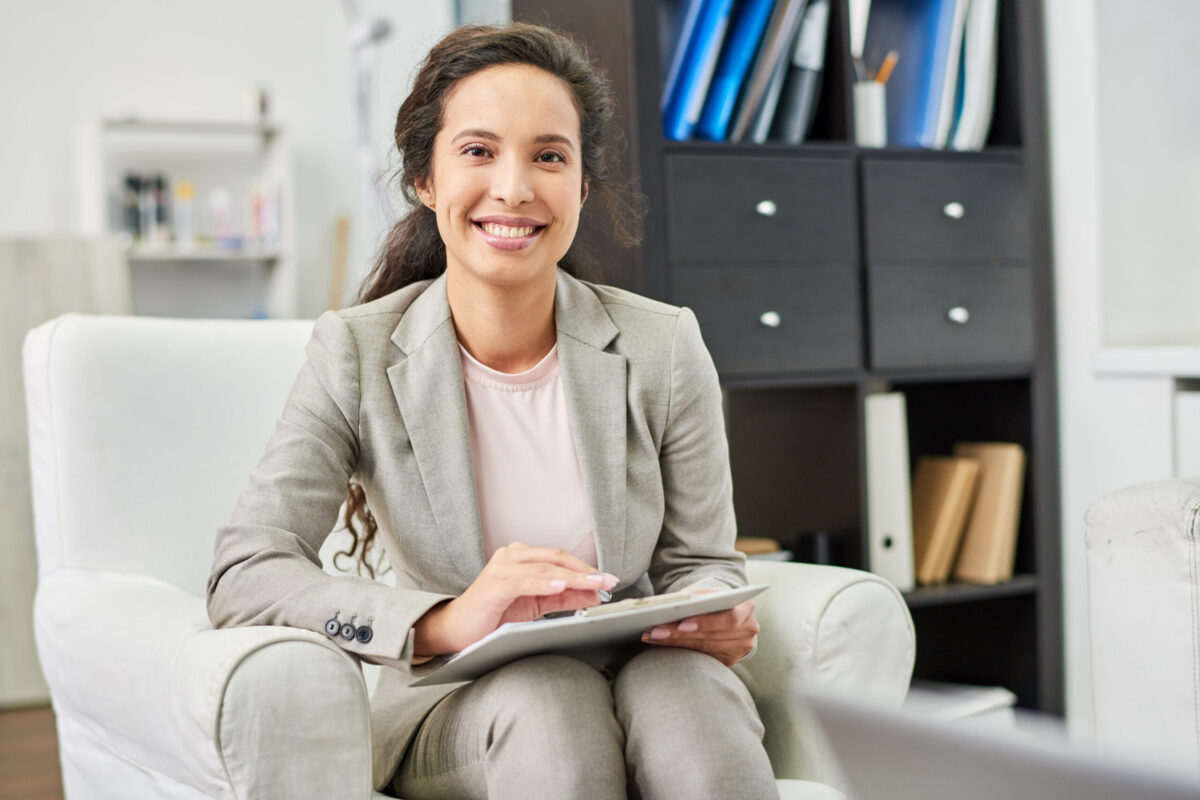 Positive successful beautiful mixed race psychologist with clipboard sitting in comfortable armchair and looking at camera in personal office